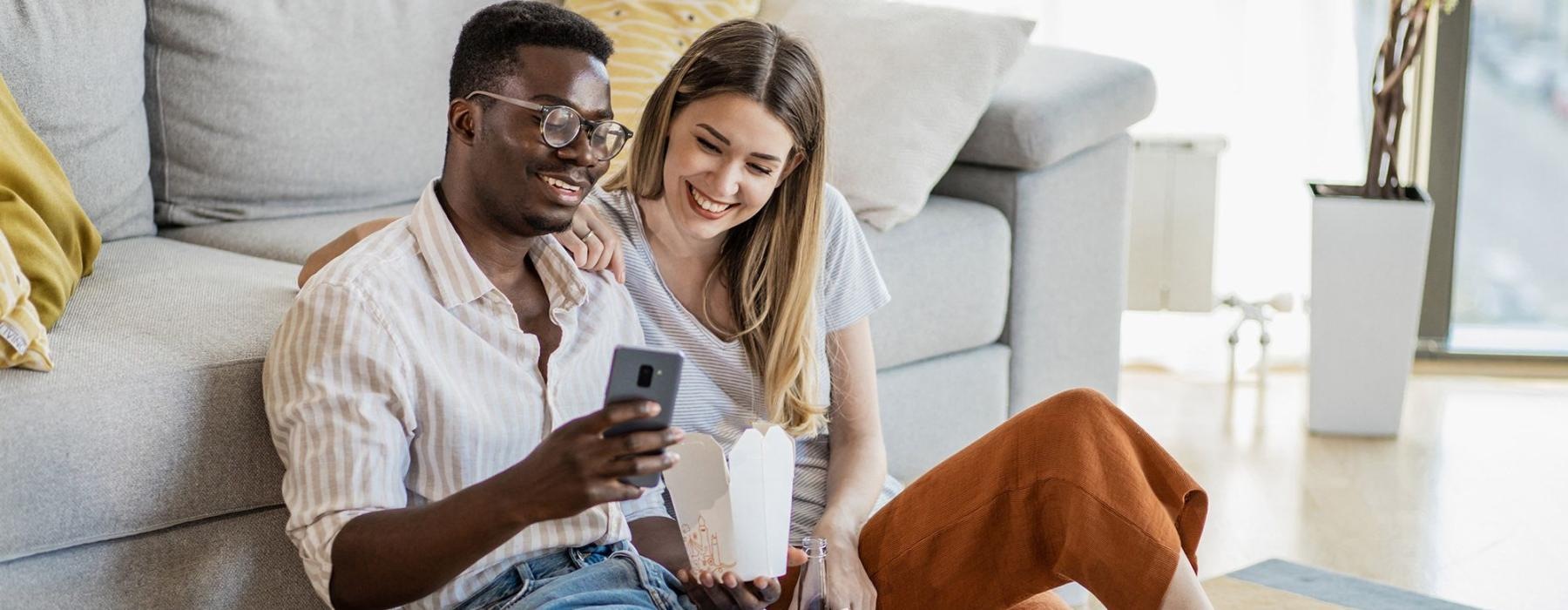 a man and woman with take out, sit against a couch on their living room floor and watch at their cell phone